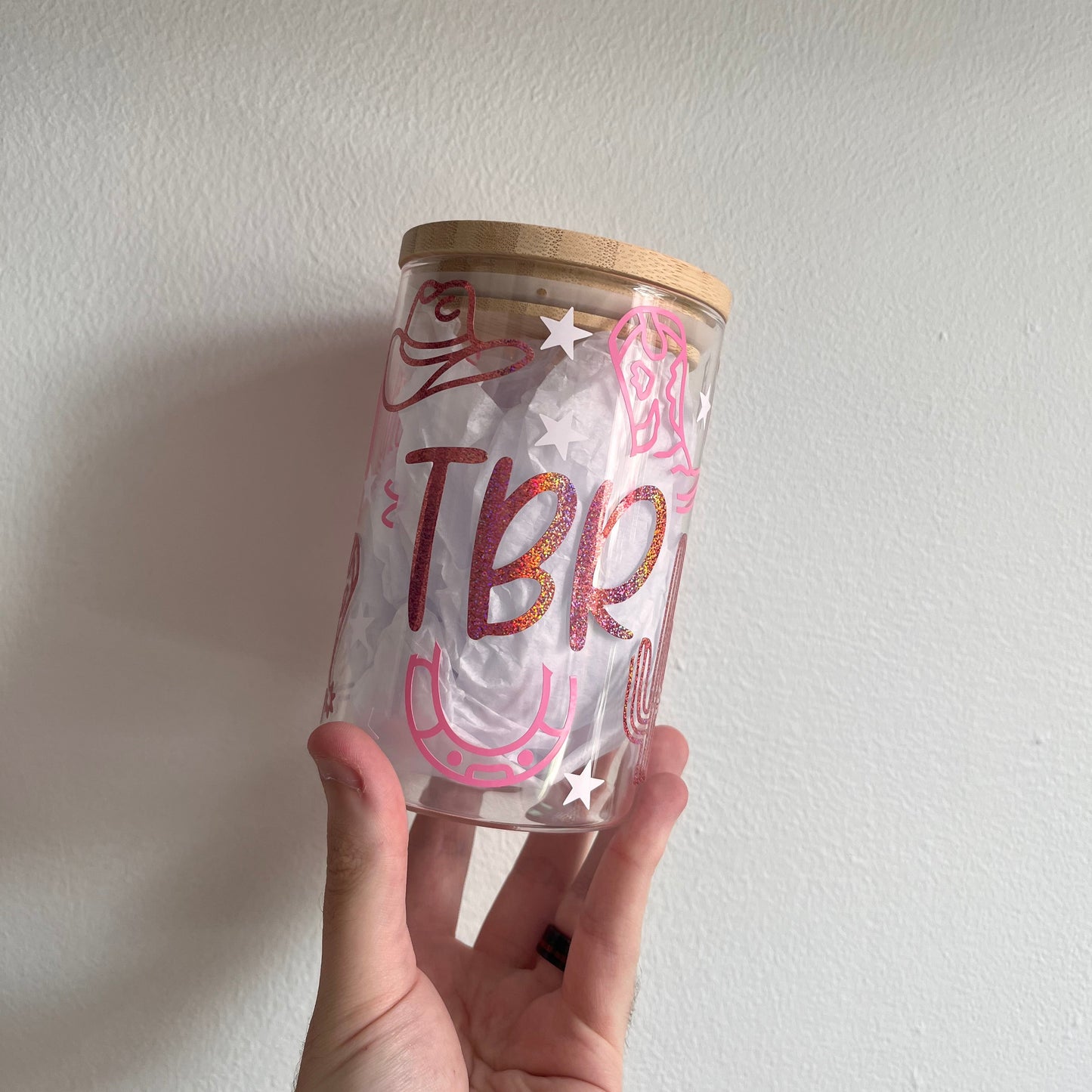 A hand holding a glass jar with a bamboo lid, decorated with pink and white vinyl text reading 'TBR' and star graphics, against a neutral background.