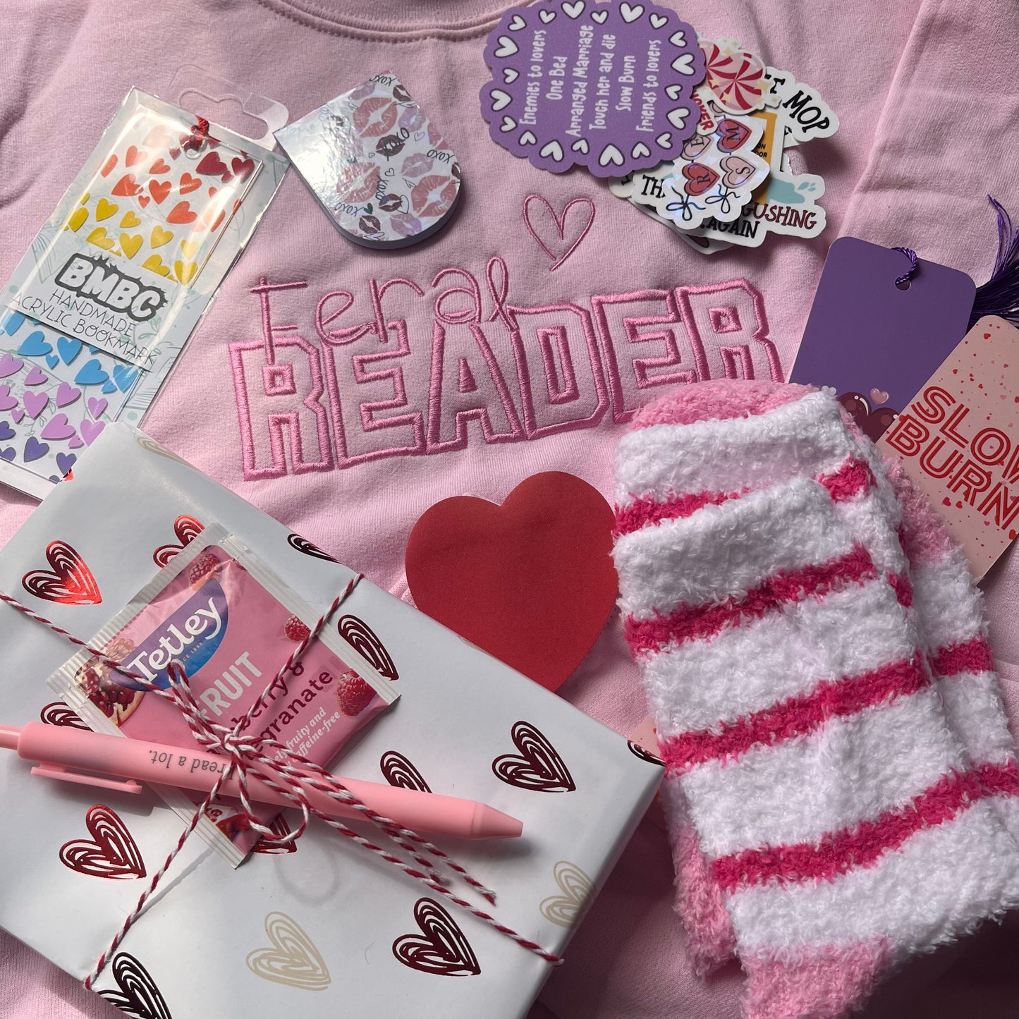 Pink t-shirt with 'Feral READER' text, colorful stickers, pink socks, and a book along with other romance themed bookish goodies