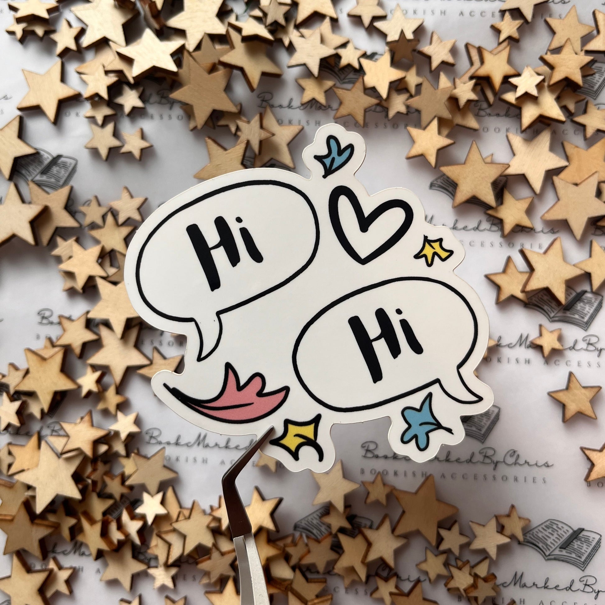 A vinyl sticker with a design consisting of two speech bubbles saying 'Hi' each other, accompanied by various simple drawings like hearts and leaves, placed on a background with multiple wooden stars.