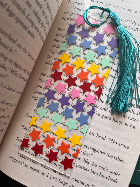 A colorful acrylic bookmark with a geometric star pattern, attached to a book page, featuring a silky soft polyester tassel.