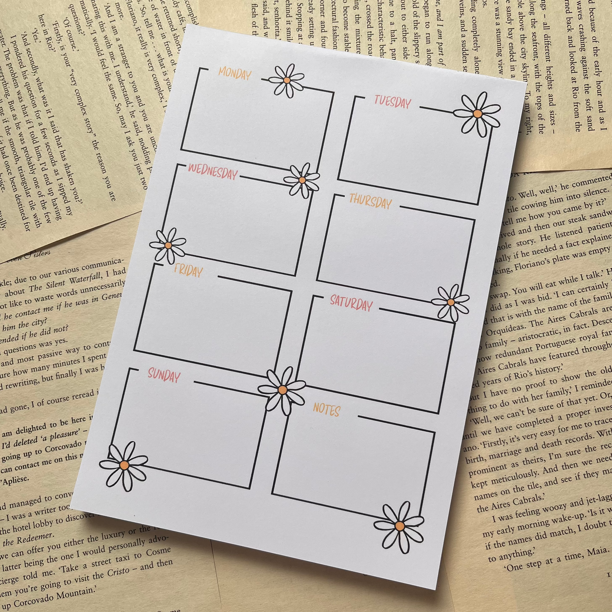 A printed sheet with a eight-panel grid, each outlined in black and decorated with a small daisy, placed on top of vintage book pages. The sheet has small peach-colored text labels inside the panels.