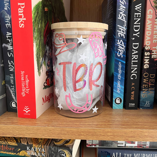 cowboy themed glass tbr jar decorated with horse shoe, boots, cowboy hats and stars with text saying "TBR" in pink and white with a bambook lid. Placed on a book shelf surrounded by books