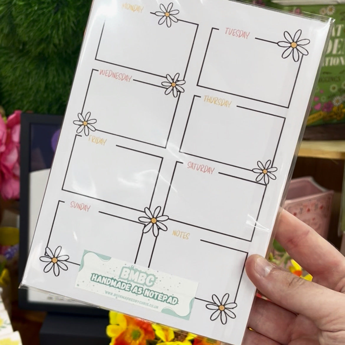 A hand holding a weekly planner notepad with daisy illustrations and labeled boxes for Monday through Sunday plus a notes section. The notepad is wrapped in clear plastic, with a store background showing flowers and shelves.