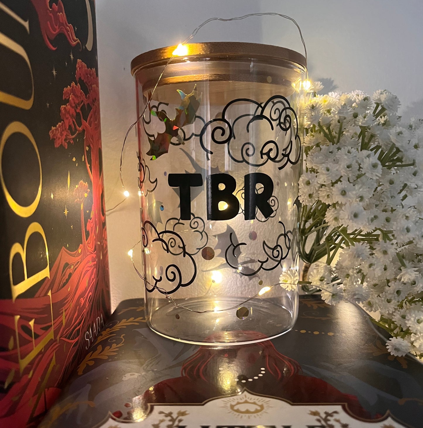 glass jar decorated in gold and black vinyl with dragons and clouds design reading "TBR" on the jar. Jar is surrounded by fairy lights, white flowers and books