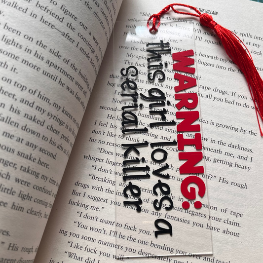 Open book with a bookmark that reads 'Warning: This girl loves a serial killer' on a green cutting mat.