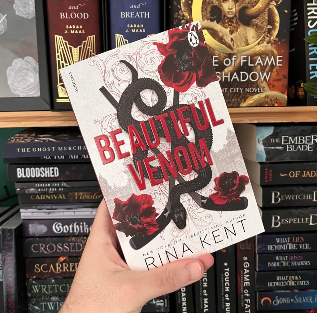 Photo shows hand holding up a book in front of bookshelves. The book held up is Beautiful Venom by Rina Kent